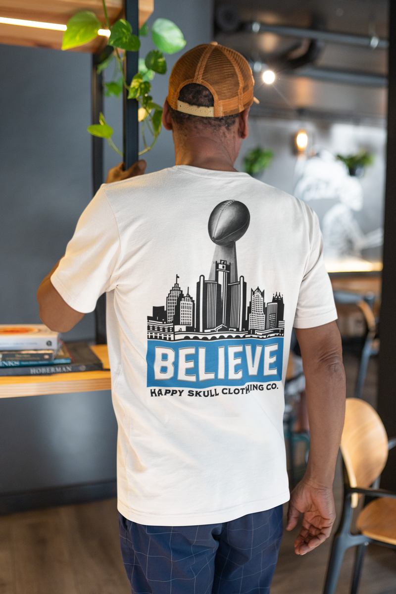 A man wearing a white t-shirt with a city skyline, a football, and the word BELIEVE printed on the back, along with Happy Skull Clothing Co. He stands indoors, touching a plant.