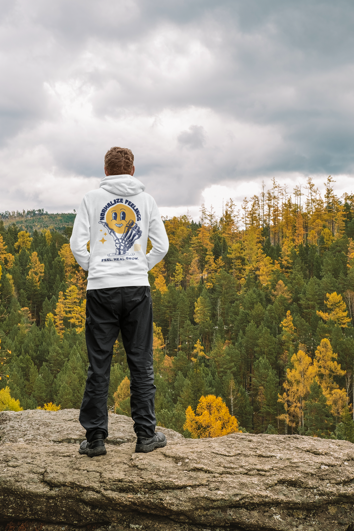 A person stands on a rocky ledge, facing a forest of green and yellow trees under a cloudy sky, wearing a white long-sleeve shirt and black pants.