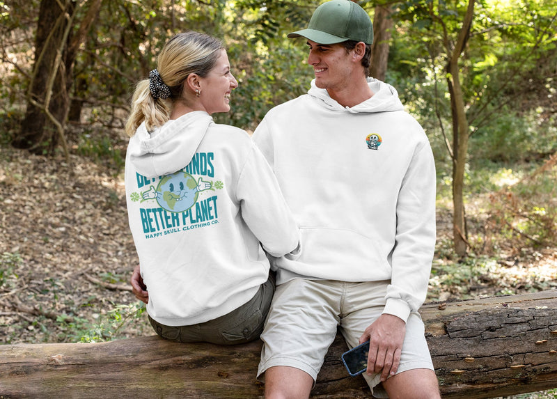 A smiling couple sits on a fallen tree in the woods. Both wear white hoodies; the woman’s hoodie has colorful earth-themed graphics and text on the back, while the man’s has a small design on the front.