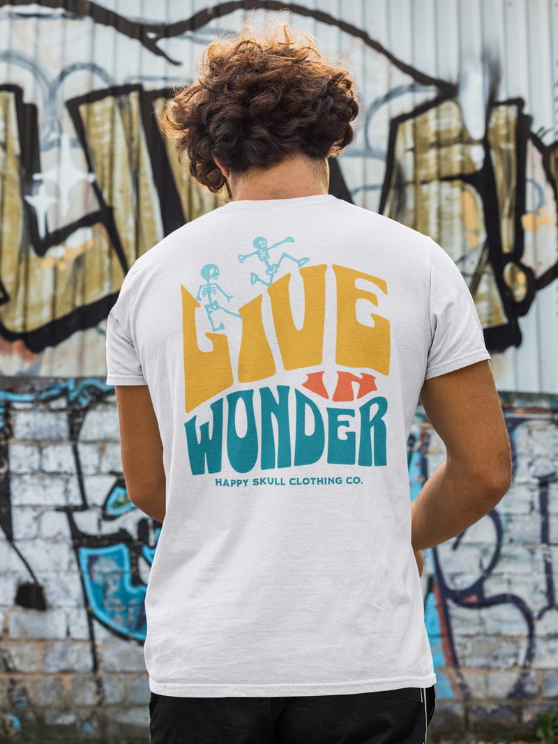 A person stands in front of a graffiti-covered wall, wearing a white t-shirt that says LIVE WONDER with cartoon skeletons and HAPPY SKULL CLOTHING CO. written underneath.