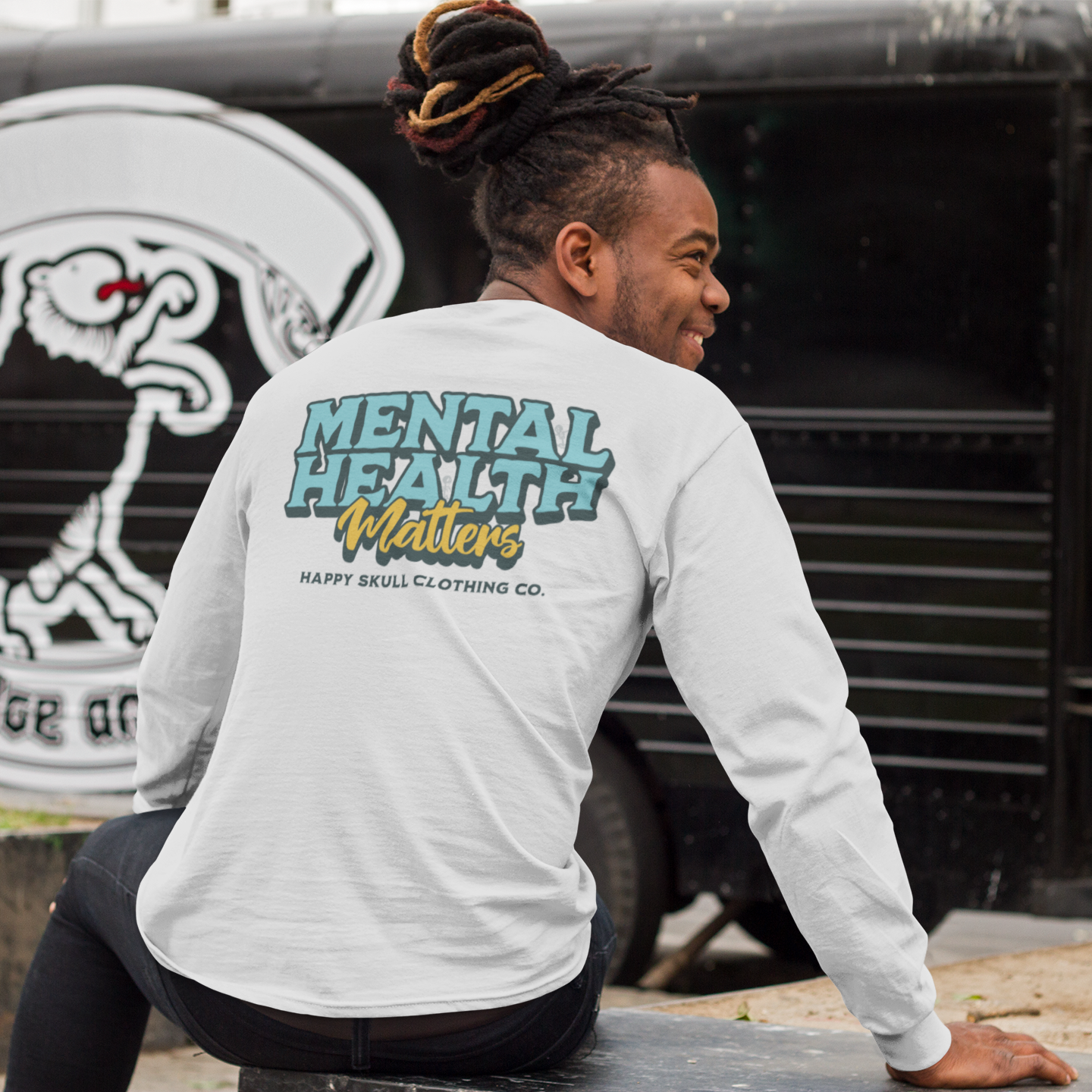 Outdoors, a smiling person with dreadlocks wears the Mental Health Matters Unisex Long Sleeve from Happy Skull Clothing Co. A black vehicle featuring a skull graphic appears in the background.
