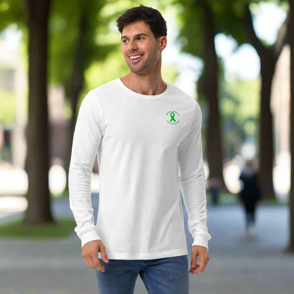A man stands smiling in a You Are Not Alone Mental Health Awareness Unisex Long Sleeve Tee, featuring a small green ribbon and text logo on the left chest. He pairs it with blue jeans against a plain black background.