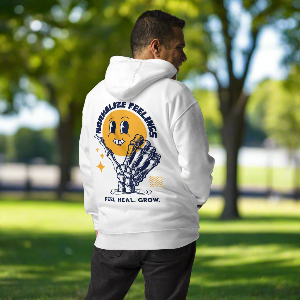 A man outdoors wears the Normalize Feelings - Unisex Hoodie, featuring a smiling yellow face and skeleton hand graphic with “Normalize Feelings” and “Feel. Heal. Grow.” text, promoting mental health among trees and grass.