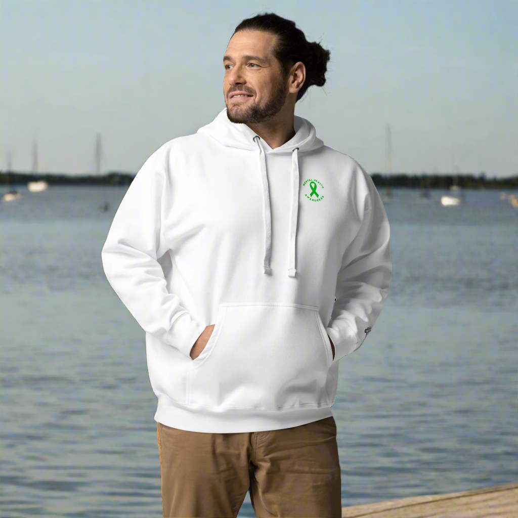A man with a beard and tied-back hair wears the You Are Not Alone Mental Health Awareness Hoodie, a white unisex top with a small green ribbon, styled with tan pants, standing hands-in-pocket against a black background.