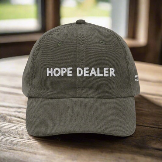 The Hope Corduroy Hat, a dark green vintage-style cap with HOPE DEALER embroidered in white on the front, rests on a wooden surface.