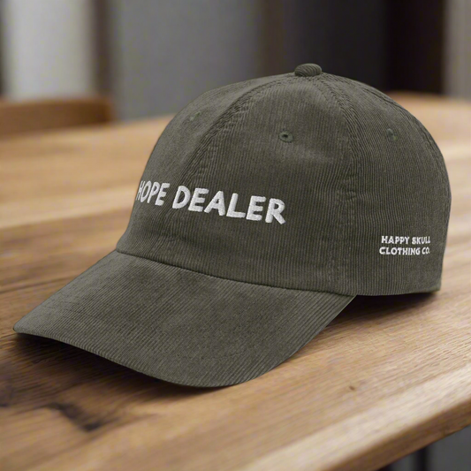 The olive green Hope Corduroy Hat features white front embroidery and HAPPY SKULL CLOTHING CO stitched on the side, displayed stylishly on a wooden surface.