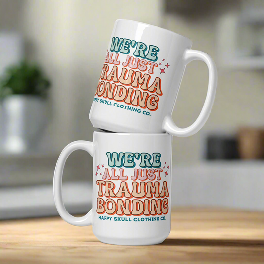Two We’re All Just Trauma Bonding - 15oz Coffee Mugs stacked on a kitchen counter, each featuring colorful text by Happy Skull Clothing Co. A fitting choice for anyone on their healing journey; the softly blurred background adds warmth.