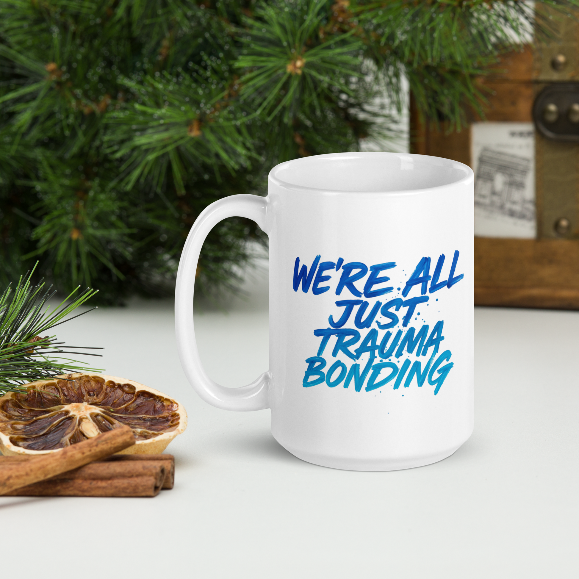 The We’re All Just Trauma Bonding 15oz Mug (New) sits on a white surface beside dried citrus, cinnamon sticks, pine branches, and a wooden box—an eye-catching ceramic mug to add to your collection.