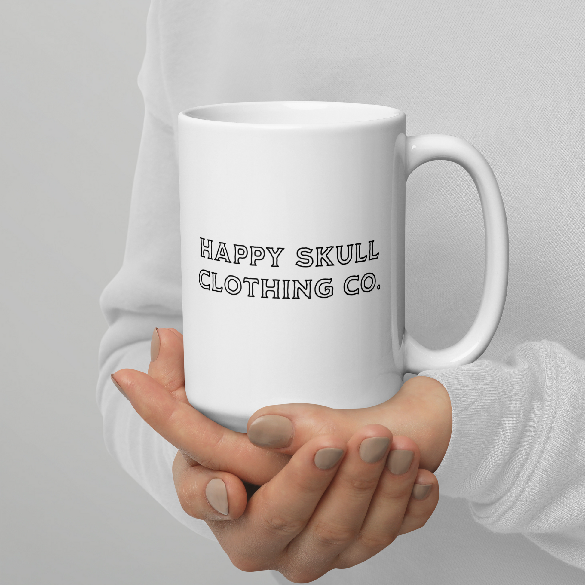 A person in a light grey sweater holds the "We’re All Just Trauma Bonding 15oz Mug (New)," a modern, dishwasher-safe ceramic mug. "HAPPY SKULL CLOTHING CO." is printed in bold black letters, and their fingernails are painted a neutral beige.