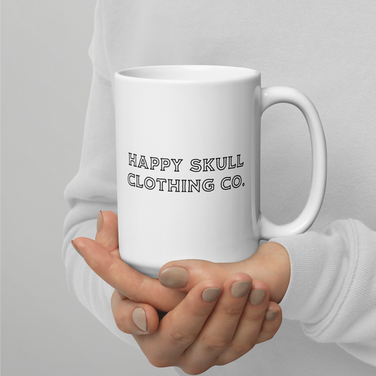 A person in a light grey sweater holds the "We’re All Just Trauma Bonding 15oz Mug (New)," a modern, dishwasher-safe ceramic mug. "HAPPY SKULL CLOTHING CO." is printed in bold black letters, and their fingernails are painted a neutral beige.